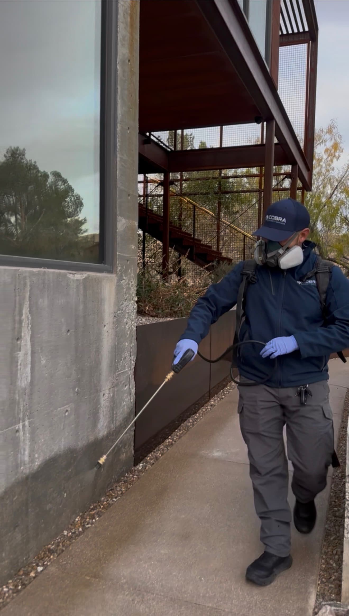 Pest control professional in protective gear sprays the exterior base of a modern concrete building.